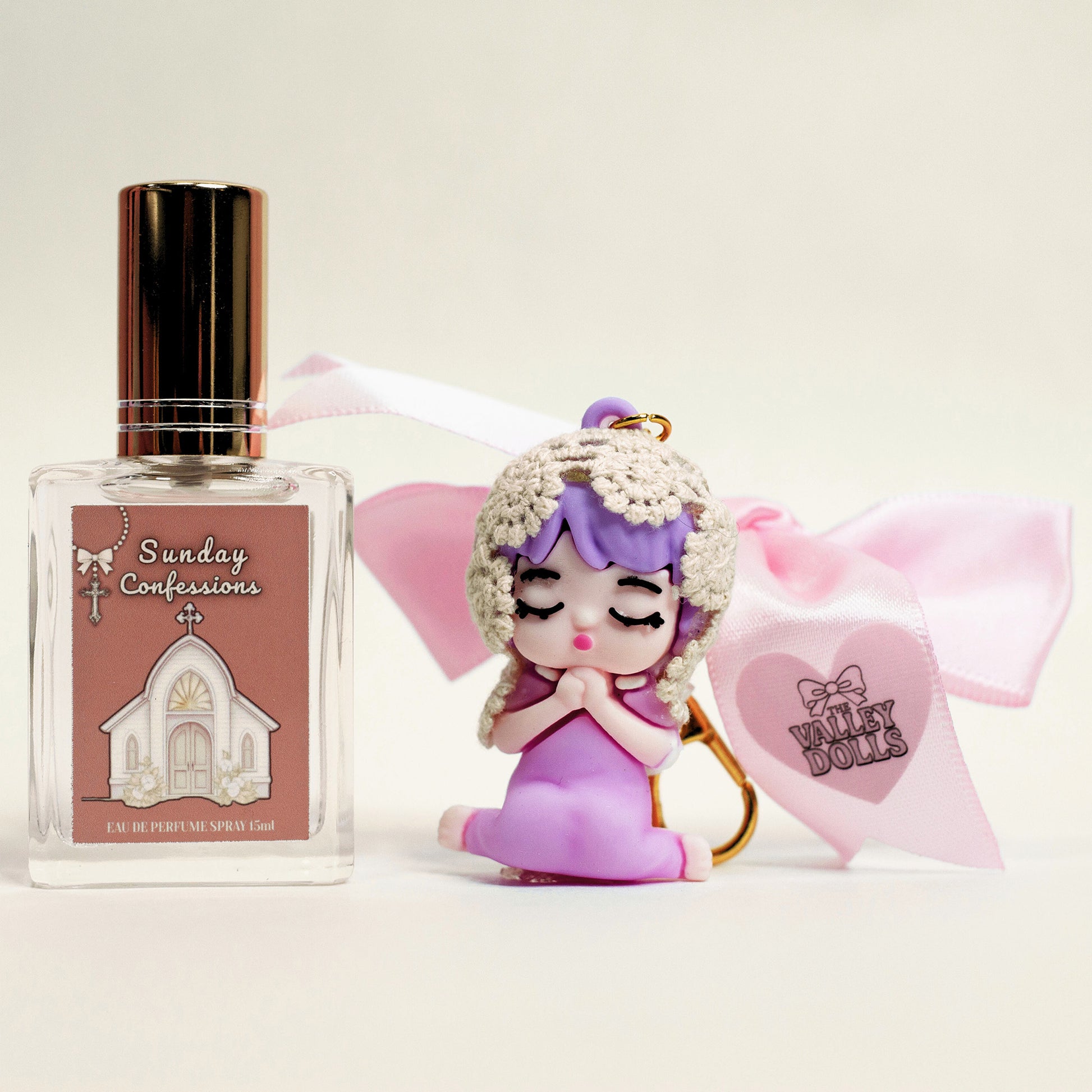 Sunday Confessions perfume spray displayed with a handmade prayer doll keychain and pink ribbon on a light background